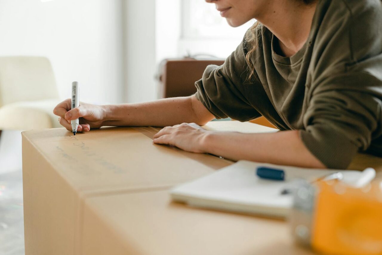 Side view of crop anonymous female in casual wear writing with marker on big cardboard box while sitting near notebook in light flat