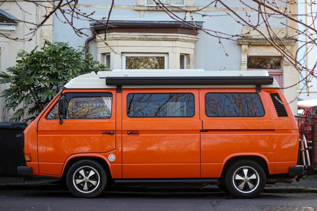 Side view of a classic orange van parked outdoors near a tree and house.